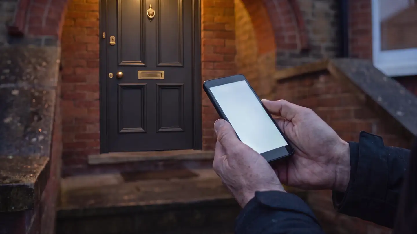 Weathered hands holding a phone with a lit, blank screen on a brick doorstep at dusk, a charcoal panelled front door behind